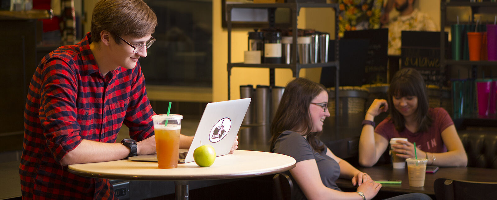 A male student uses a laptop while standing at a table in the 1855 Room with a drink and apple while two female students with drinks talk in the background while sitting at another table.