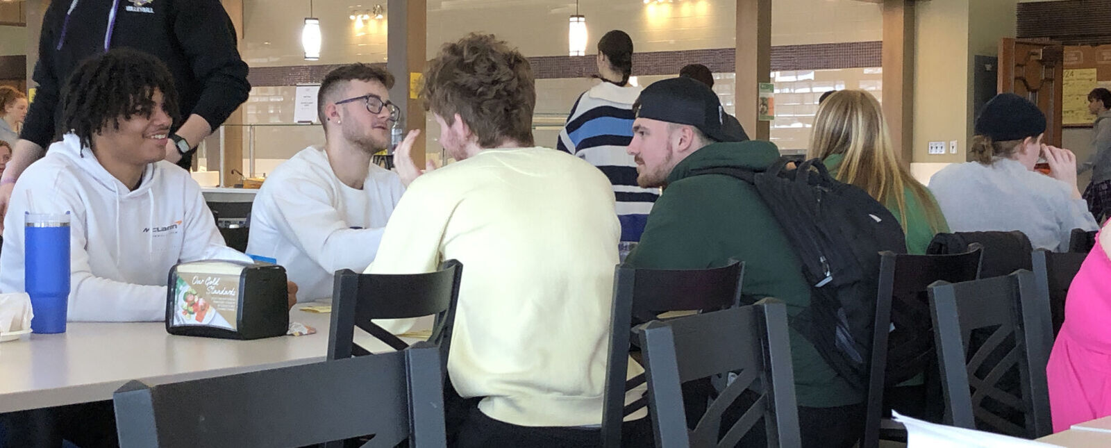 A group of students talk and laugh around a dining hall table.