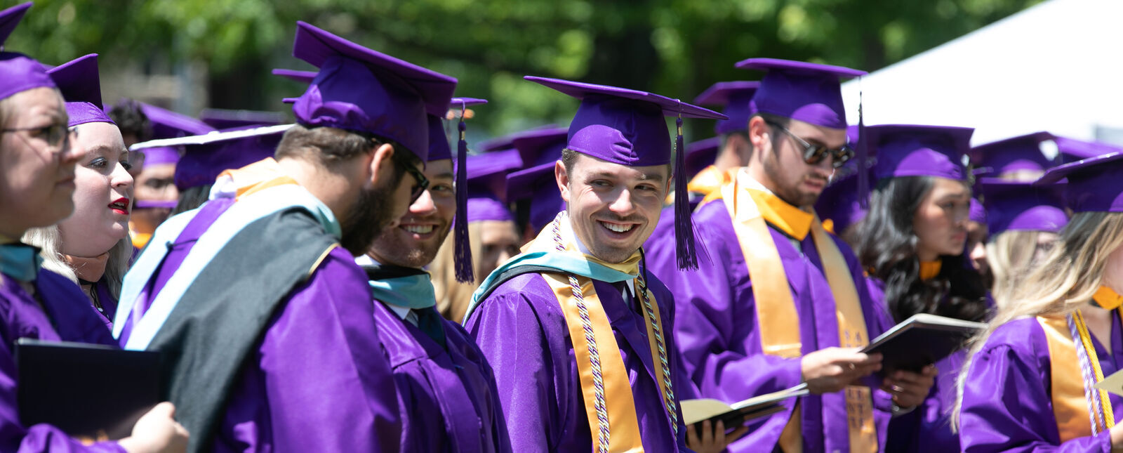 Some of the graduates smile at each other while standing during part of Commencement