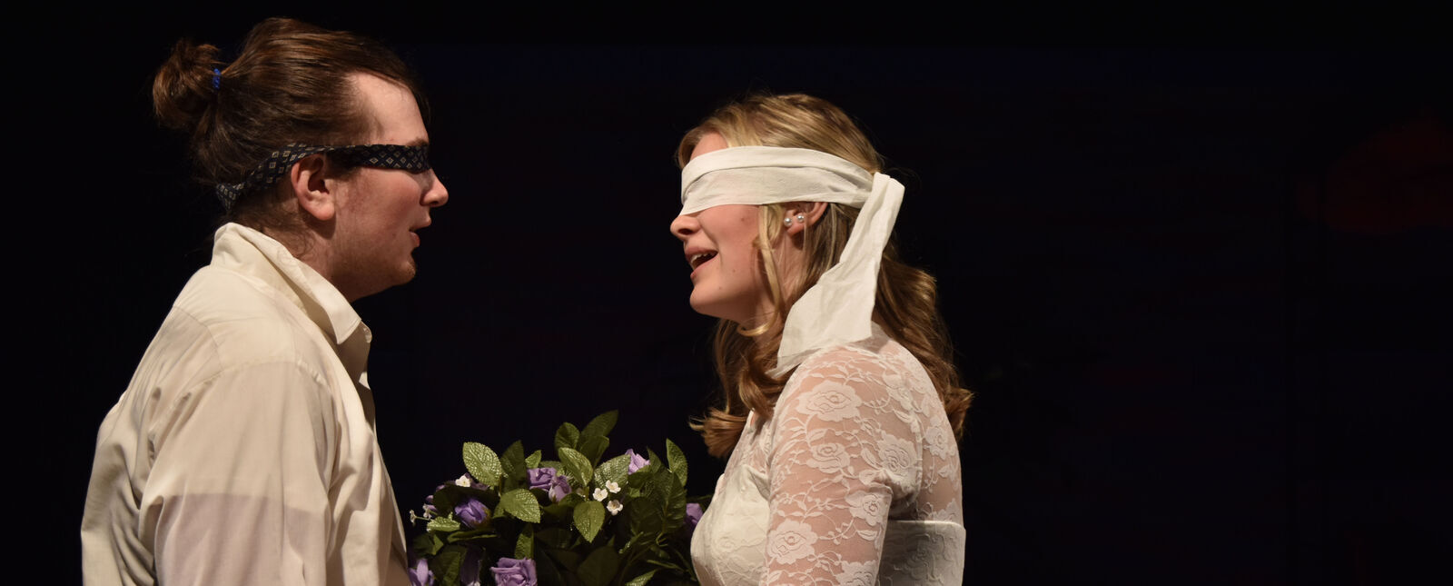 A male and female student wear blindfolds and face each other wearing wedding attire in the play Love/Sick