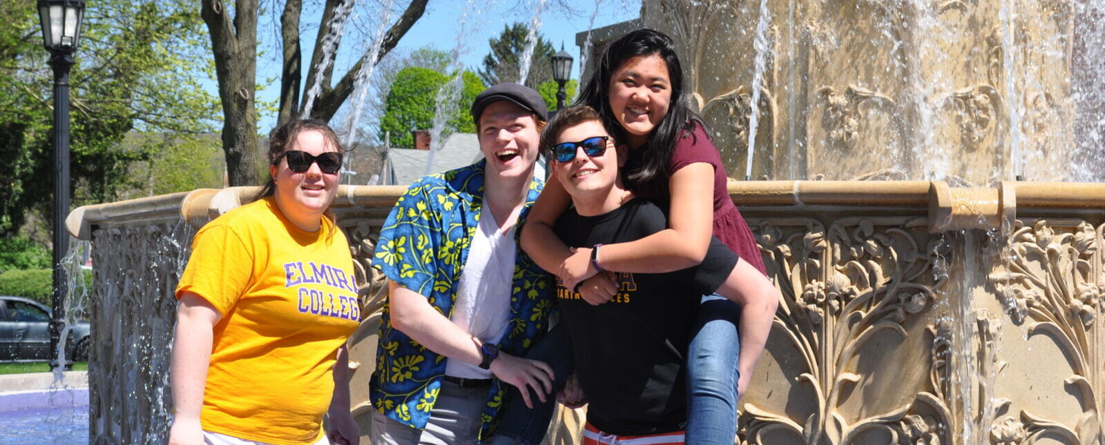 Four Elmira College alumni, one riding piggy back, smile for a picture in the fountain