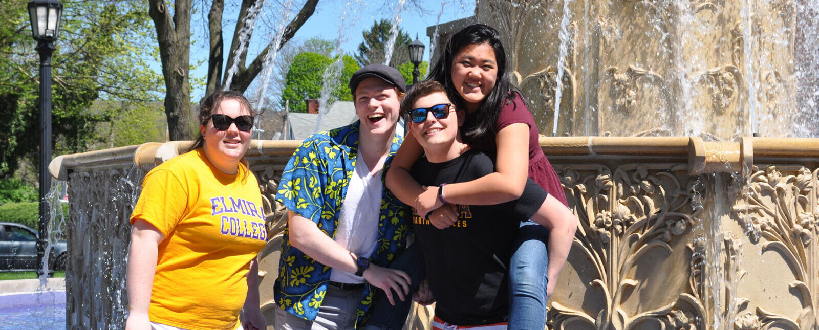 Four Elmira College alumni, one riding piggy back, smile for a picture in the fountain