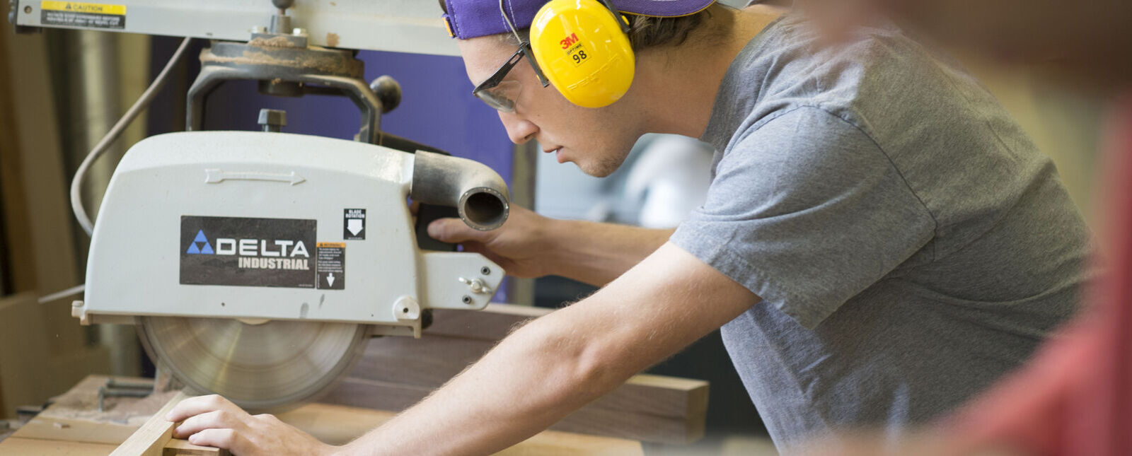 A male student wearing ear protection cuts into a piece of wood using a table saw.