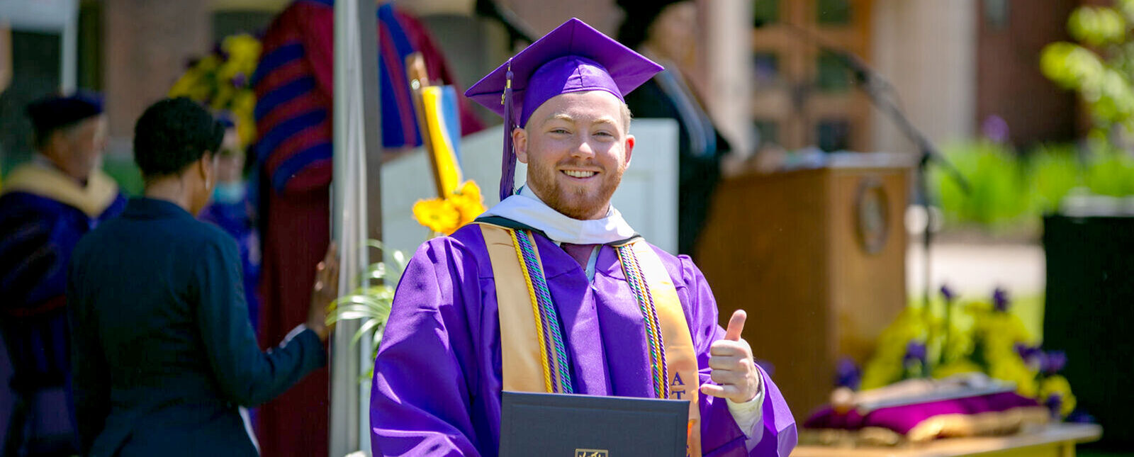 A male graduate gives a thumbs up to the camera after receiving his degree