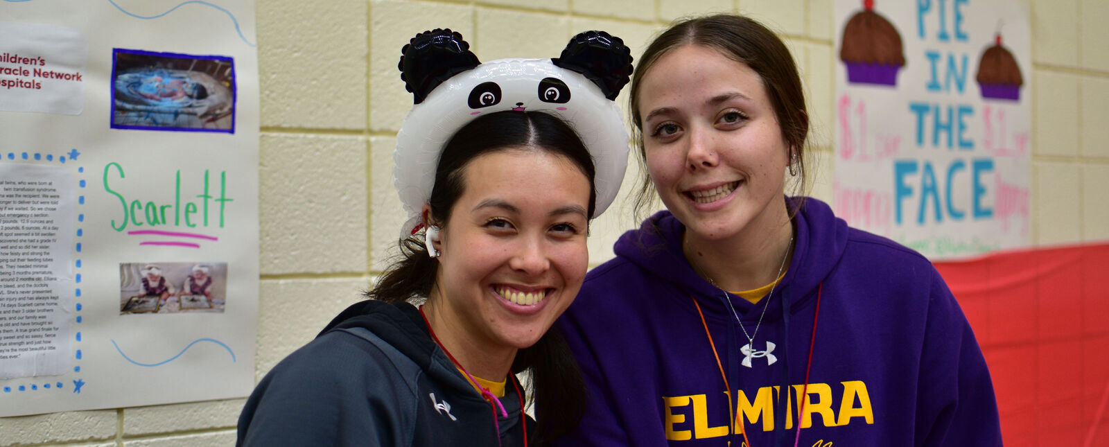 Two female students – one wearing a blow up panda hat – smile together during the Elmira College Dance Marathon in Speidel Gymnasium