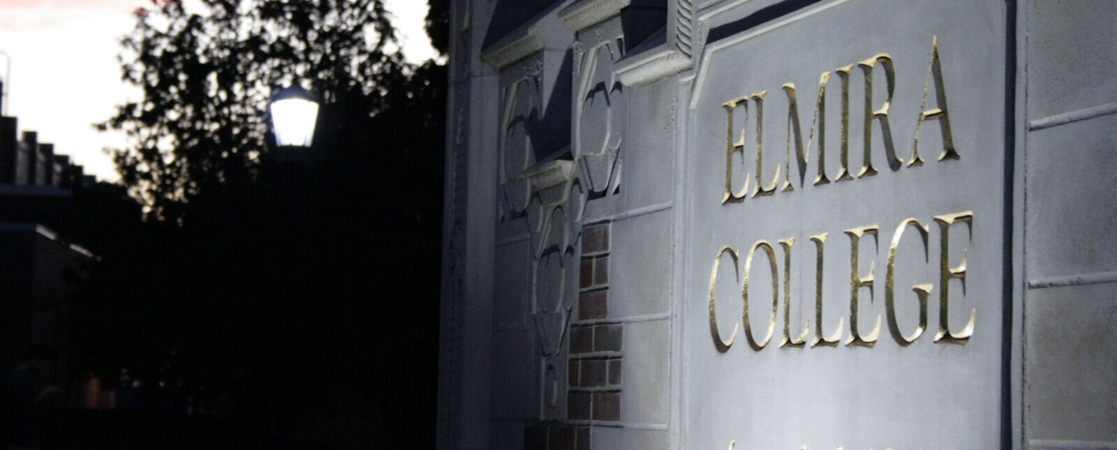 A stone Elmira College sign is lit up by spotlights during dusk