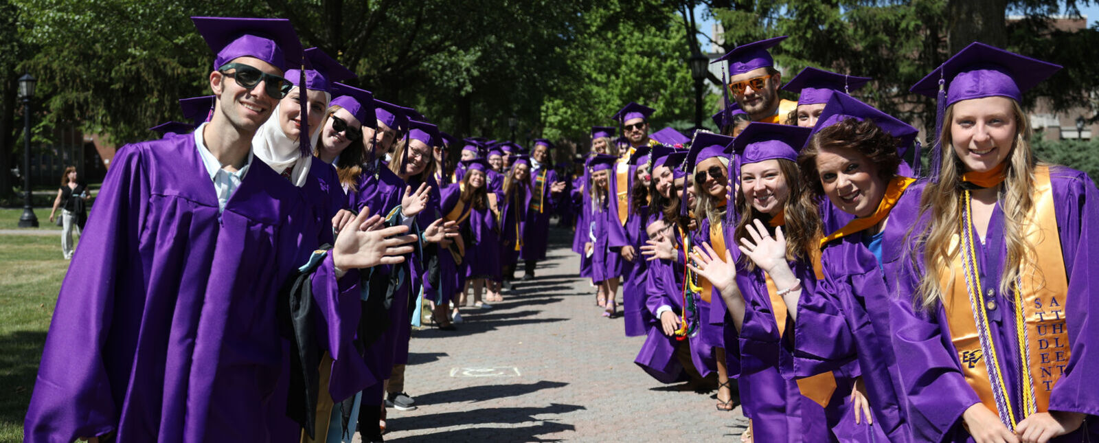 Graduates lined up on the sides of a walkway wave to the camera
