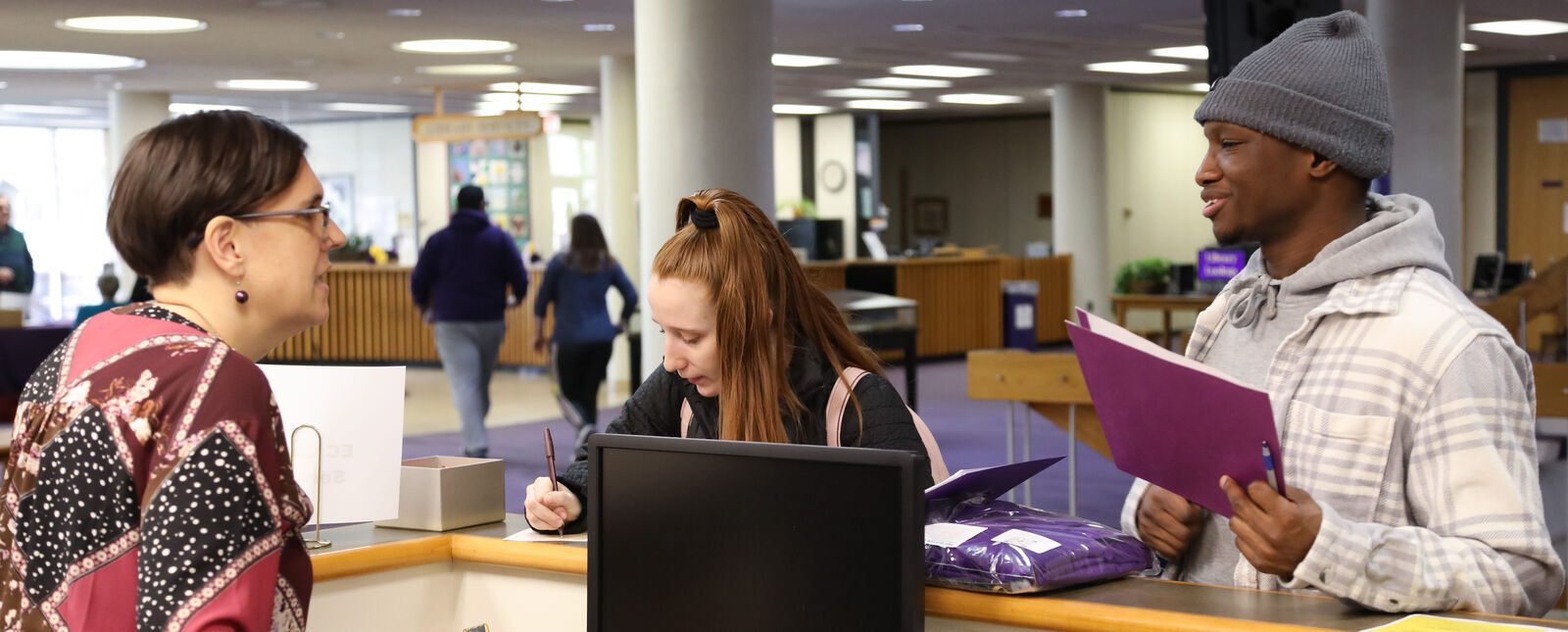 A male student talks with a Career Services employee as a female student fills out a form on the counter of the Career Services desk in the Gannett-Tripp Library