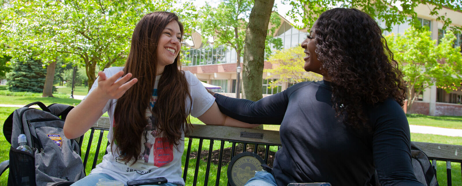 Two female students laugh while talking on a bench