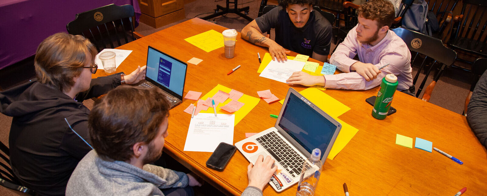 Students work together on laptops and with papers around a wooden table