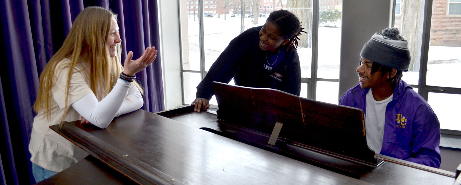 A female student and two male students share some laughs around a piano in the Towers residence hall.