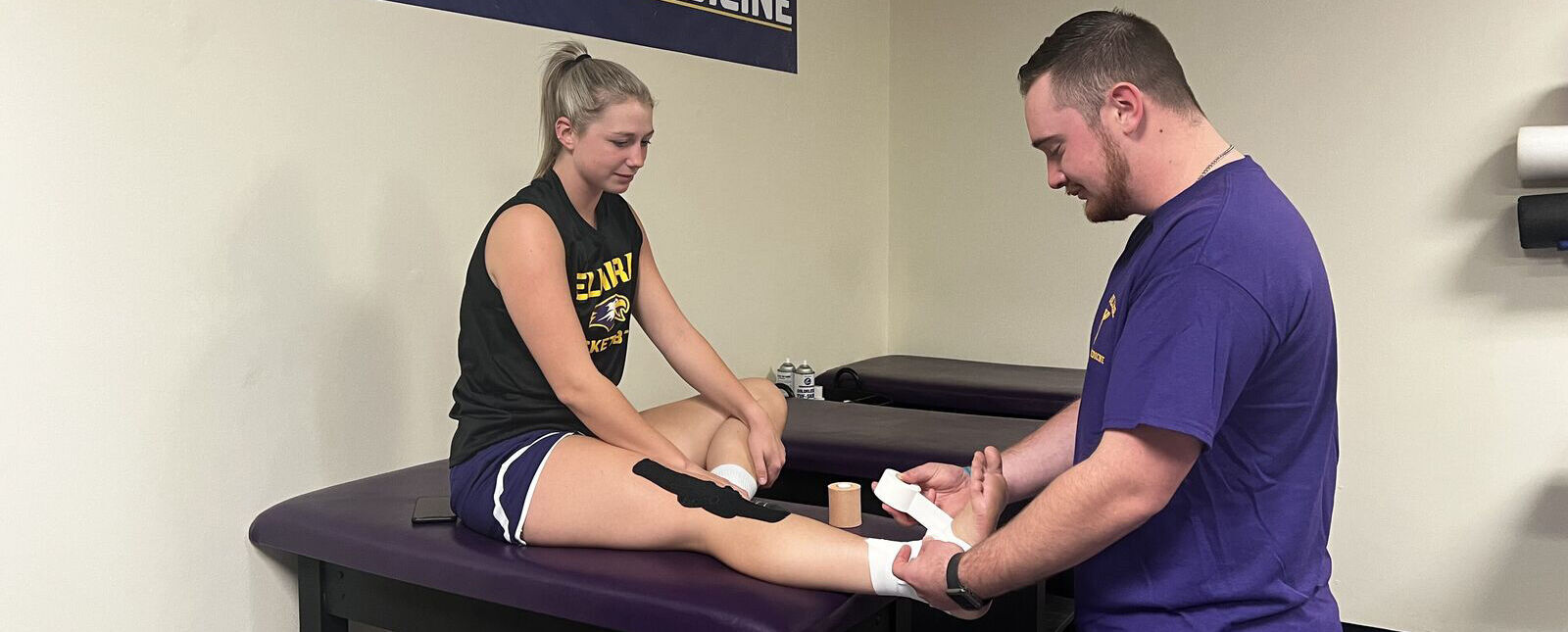 A male trainer tapes up the ankle of a female student athlete