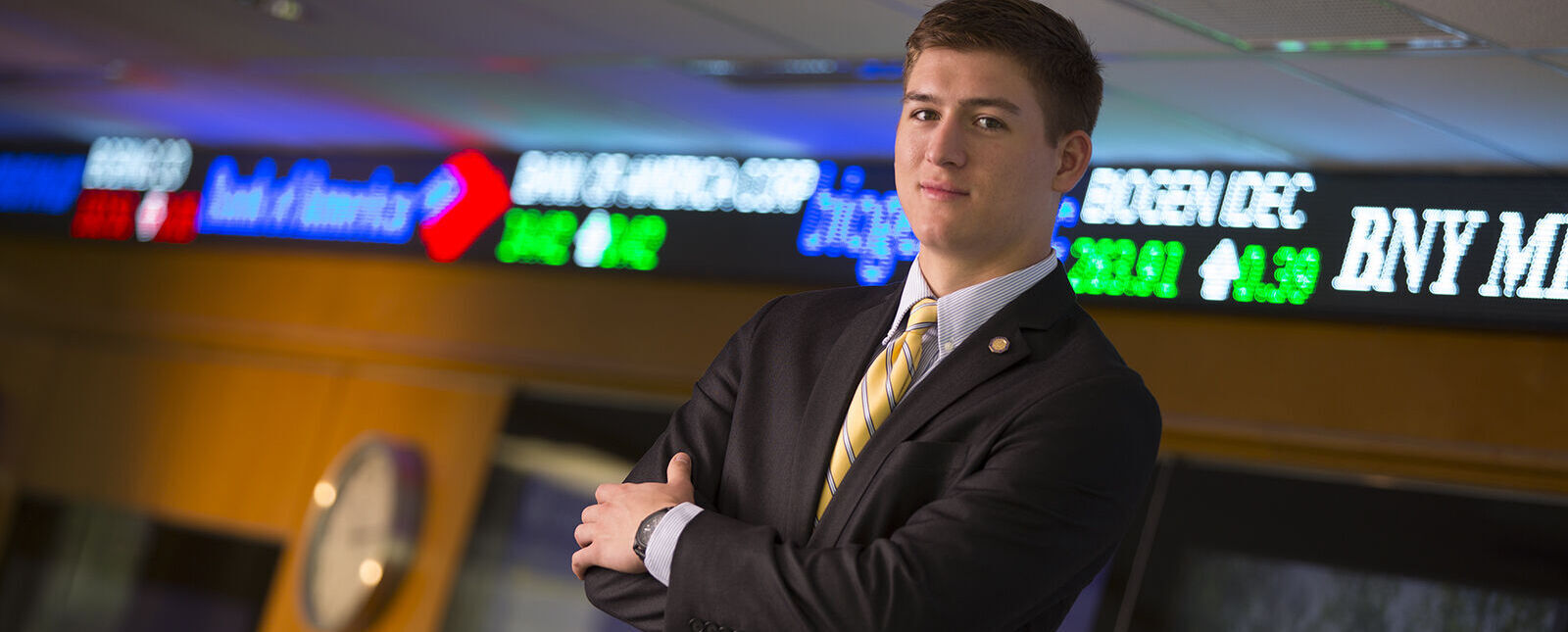 A male student poses in a business suit with arms crossed in the Malesardi Finance Trading Room