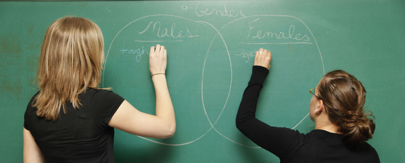 Two female students start lists about males and females on a chalkboard